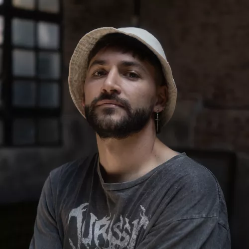 El Fran mirando a la cámara con expresión seria, vistiendo remera gris desgastada y sombrero tipo pescador beige. De fondo, se distingue una pared de ladrillo y una ventana de vidrio repartido desenfocada.
