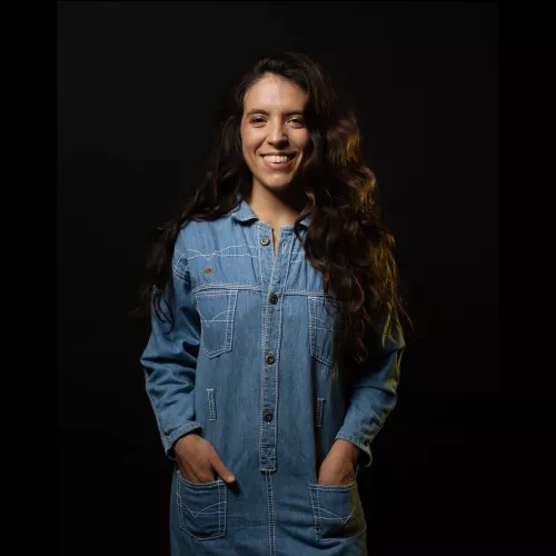 Mujer sonriente, de cabello largo, abundante y ondulado. Vestida con un enterito de jean ochentoso