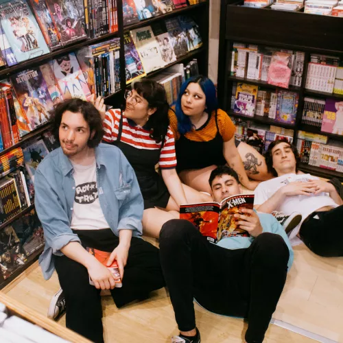 Un grupo de 2 chicas y 3 chicos sentados en el piso de un local de historietas leyendo y mirando cómics