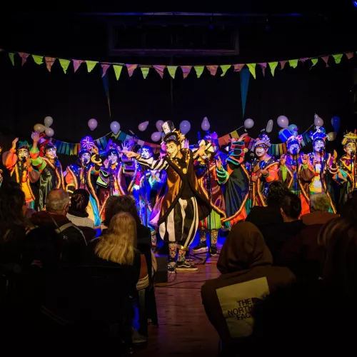 se visualiza a la murga con sus vestuarios de colores llamativos y los rostros pintados con maquillaje artístico. Están en un teatro finalizando un acorde.