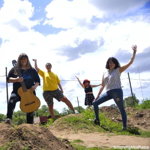 Las cuatro saludando. Laura tiene una guitarra en la mano. Es un día claro con nubes. Estamos paradas sobre una loma de tierra y pasto.