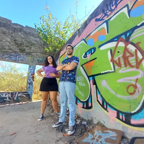 loca la calandria: "Fiore y Santi posan en un espacio urbano abandonado, junto a una pared con grafitis. La mujer lleva una camiseta morada y falda negra, mientras que el hombre viste una camisa estampada y jeans. La escena está iluminada por la luz del día, destacando el contraste entre la vegetación que se cuela por las ventanas sin vidrio y los coloridos grafitis en la pared.