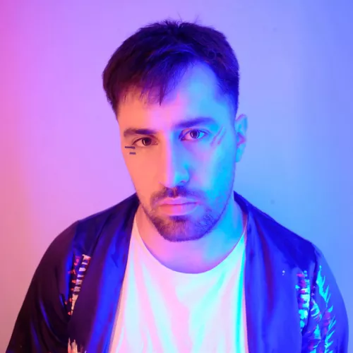 Foto de plano medio corto, colores rosas y celeste tiñen el fondo, Manu Elle mira de frente a la camara