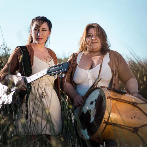 Mariel y Noelia estan con guitarra y bombo en mano entre juncos junto al rio