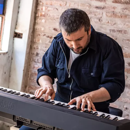 Ramiro Santos tocando un teclado con concentración junto a una ventana en un ambiente de ladrillo a la vista.