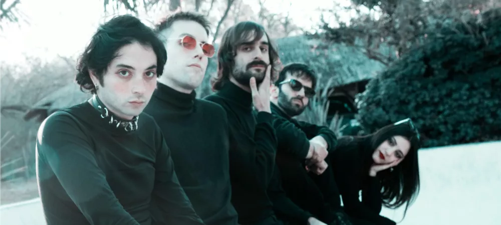 Los 5 integrantes sentados al borde de una piscina retro, vestidos de negro, miran a camara en hilera.