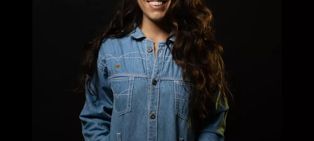 Mujer sonriente, de cabello largo, abundante y ondulado. Vestida con un enterito de jean ochentoso
