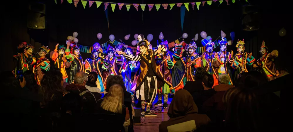 se visualiza a la murga con sus vestuarios de colores llamativos y los rostros pintados con maquillaje artístico. Están en un teatro finalizando un acorde. 