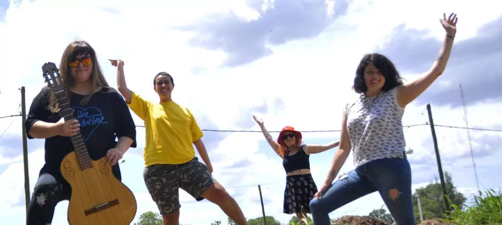 Las cuatro saludando. Laura tiene una guitarra en la mano. Es un día claro con nubes. Estamos paradas sobre una loma de tierra y pasto.