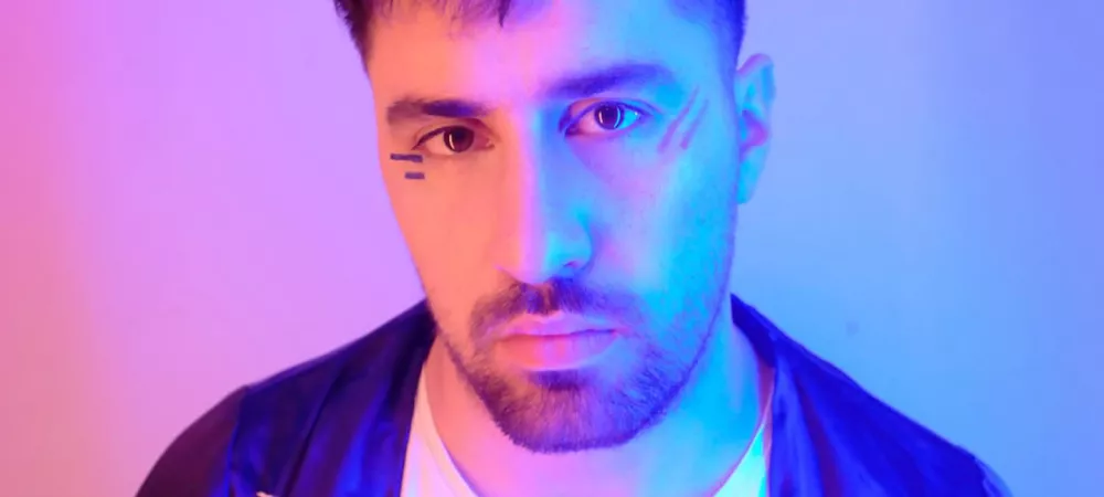 Foto de plano medio corto, colores rosas y celeste tiñen el fondo, Manu Elle mira de frente a la camara