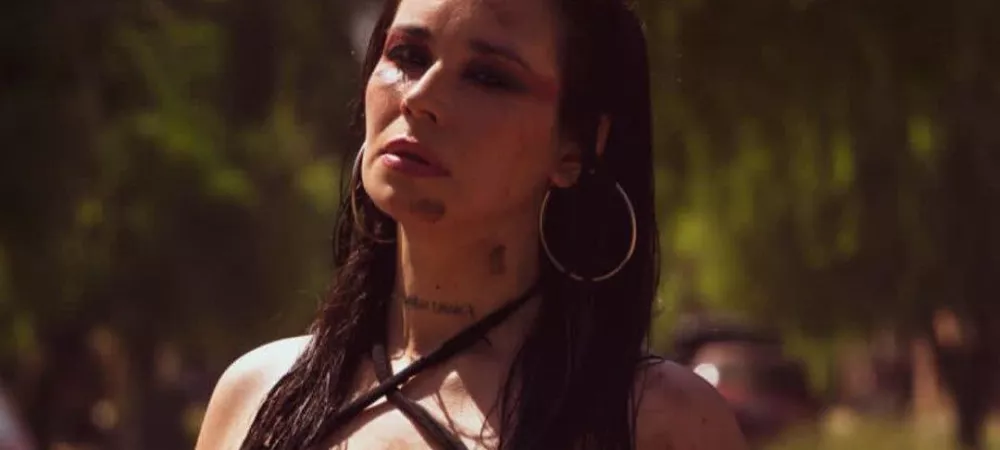 Se observa mujer de cabello largo negro al aire libre, de fondo un barrio bonaerense, maquillada y musculosa cruzada en el pecho negra con tachas diradas. Su rostro expresa suciedad y calor sofocante.