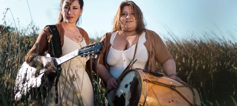 Mariel y Noelia estan con guitarra y bombo en mano entre juncos junto al rio