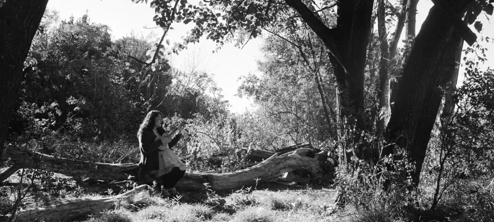 se visualiza a la artista sentada en un tronco en un espacio similar a un bosque, tocando el ukelele