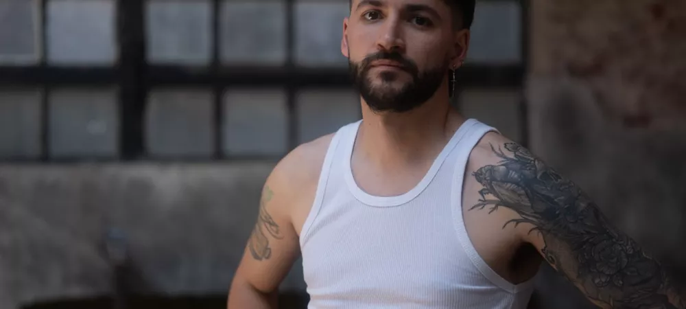 El Fran de pie en un ambiente interior con luz natural suave, vistiendo remera sin mangas blanca y jeans. Tiene tatuajes en ambos brazos y mira a la cámara con expresión seria. Al fondo, desenfocada, se ve una guitarra apoyada sobre un sillón verde frente a una gran ventana de vidrio repartido.