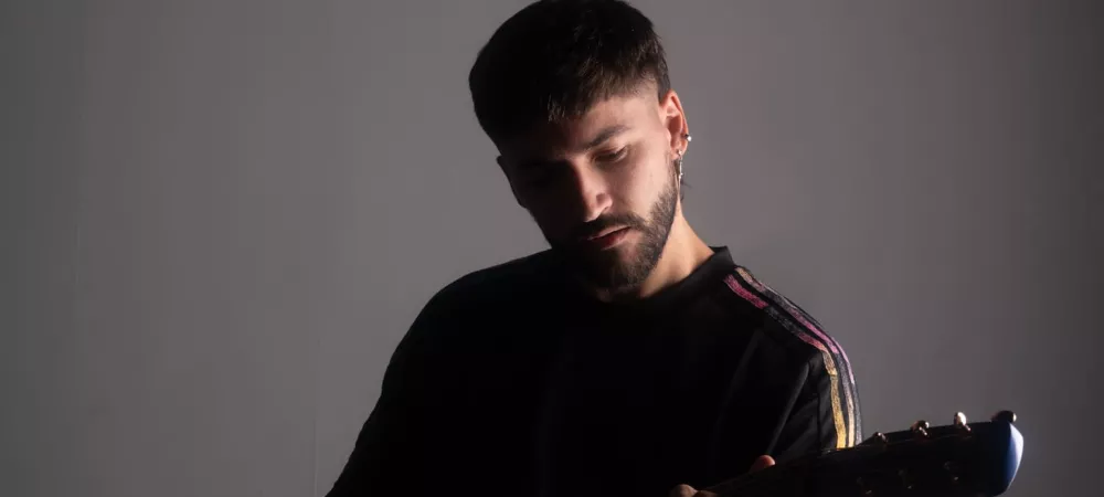 El Fran tocando una guitarra azul en un ambiente con fondo gris liso. Viste remera negra con franjas de colores en las mangas y luce concentrado, mirando hacia el instrumento bajo una iluminación tenue que resalta su silueta.