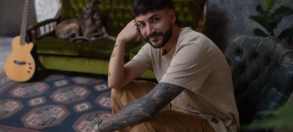 El Fran sentado en una silla verde gastada, apoyando la cabeza en su mano y sonriendo a la cámara. Viste remera beige y pantalón marrón claro. Al fondo, sobre un sillón verde, hay un perro recostado y una guitarra apoyada, en un ambiente con pared rústica, gran ventana y alfombra decorativa.
