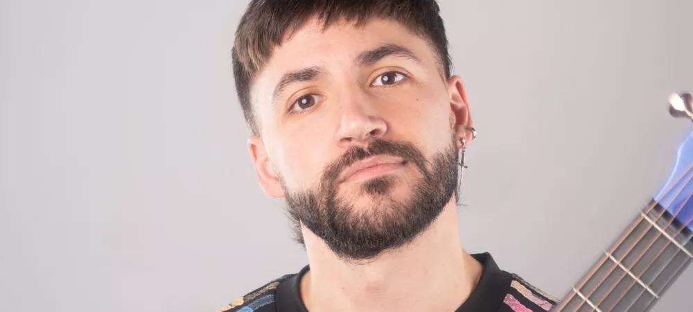 El Fran sosteniendo una guitarra azul en posición vertical frente a su pecho, mirando a la cámara con expresión serena. Viste remera negra con franjas de colores en las mangas, sobre un fondo gris claro.