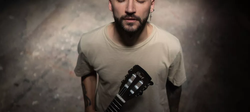 El Fran de pie en un espacio oscuro, iluminado por un haz de luz cenital. Viste remera beige y sostiene una guitarra marrón en posición vertical, con los ojos cerrados y expresión serena, transmitiendo una sensación de introspección.