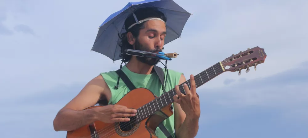 Cielo. Adelante un hombre con un paraguas en la cabeza, con una armonica en la boca, tocando una guitarra criolla