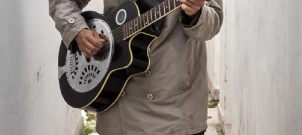 Un pasillo, adelante un hombre parado, con saco, el pelo le tapa la cara, toca una guitarra de metal