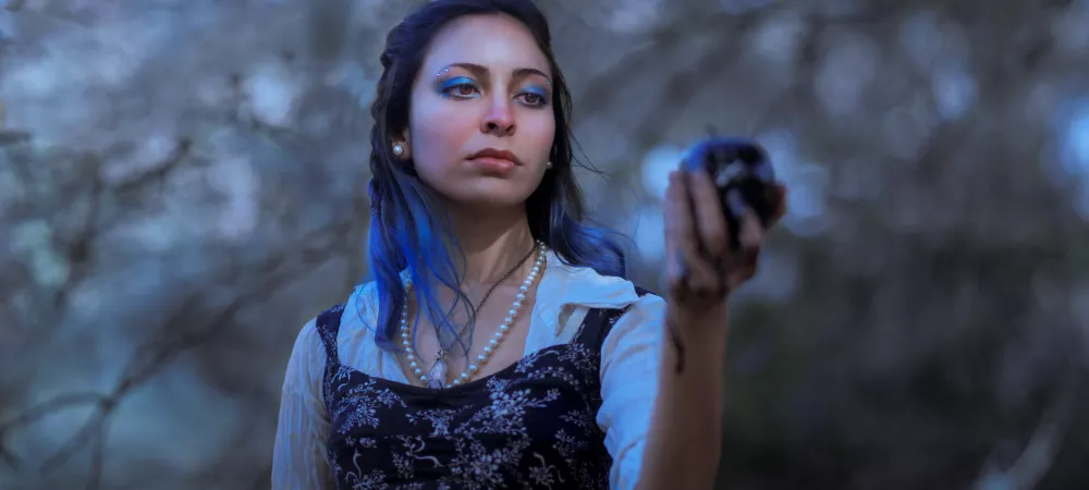 Imagen de una mujer joven de pelo azul con mirada triste mirando una manzana negra que sostiene en su mano