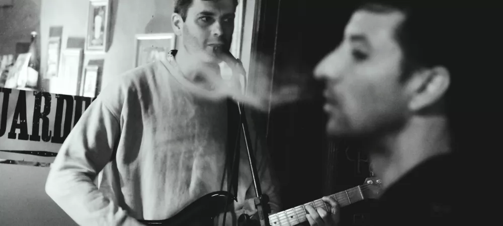 Em músico tocando y en frente un hombre fumando de perfil. De fondo se ve la vidriera de un bar