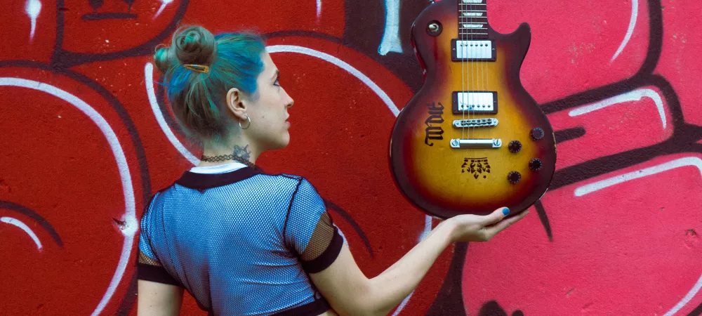 La imagen muestra a la artista Jsefina sosteniendo su guitarra en una de sus manos, alzandola mientras la observa