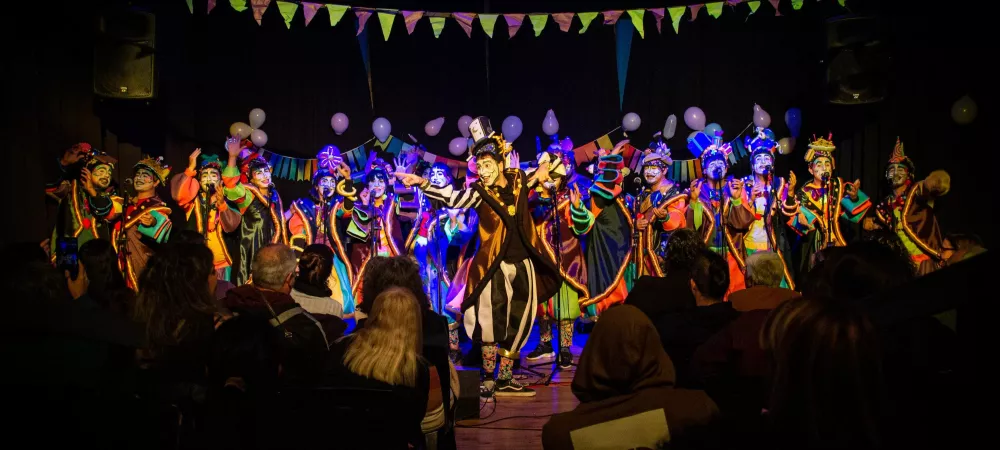 se visualiza a la murga con sus vestuarios de colores llamativos y los rostros pintados con maquillaje artístico. Están en un teatro finalizando un acorde. 