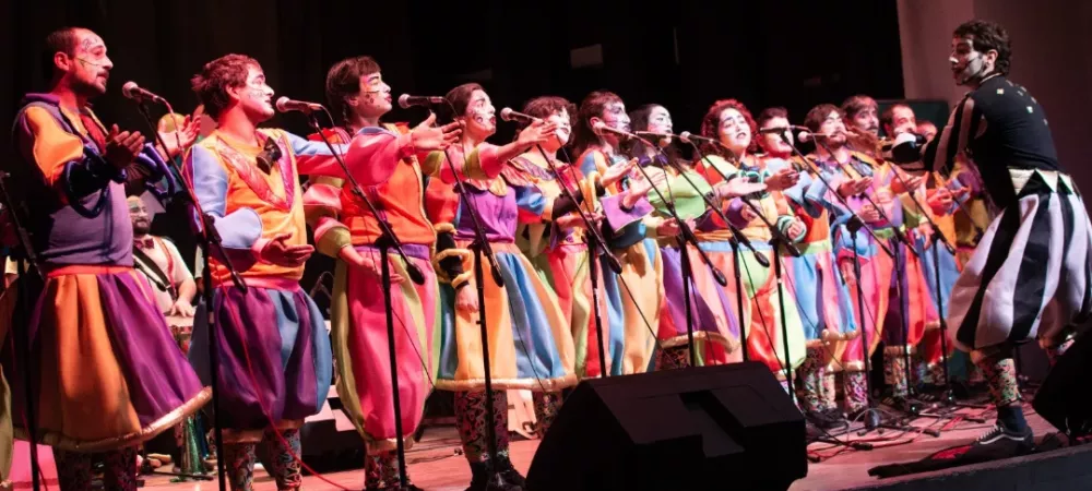 se visualiza al coro de la murga con trajes coloridos cantando arriba de un escenario