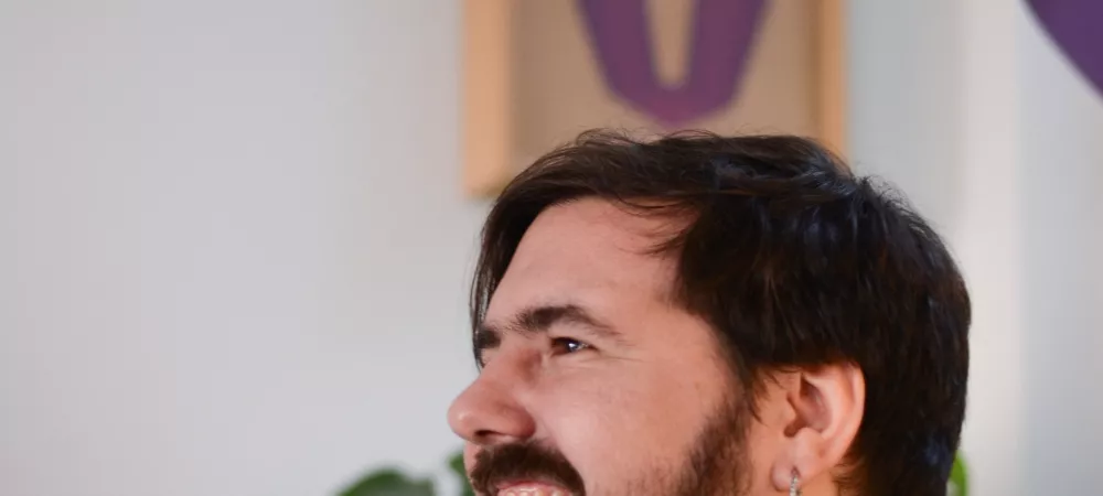 Yo de perfil. Atras de mi una planta, un cuadro sobre una pared blanca y una tela de colores colgando