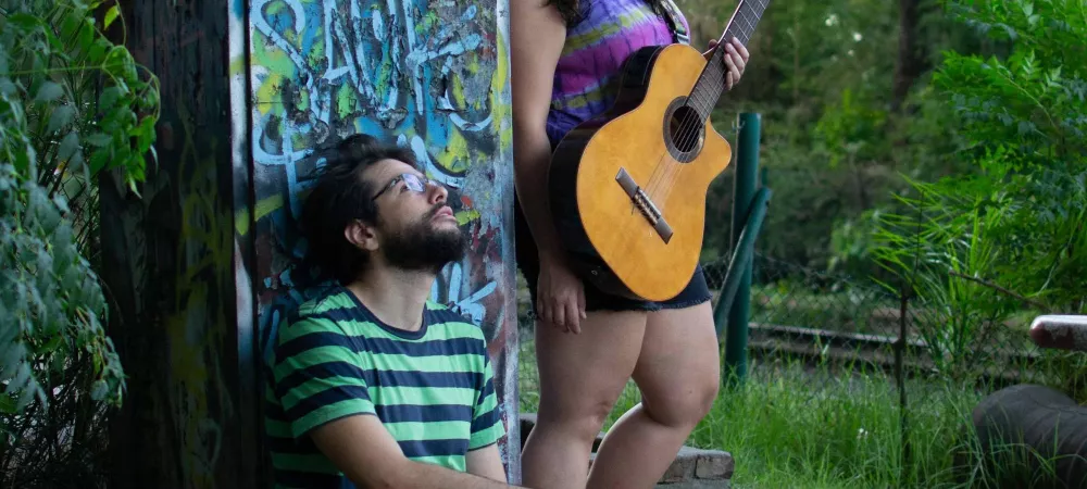 Dos músicos posan en un entorno al aire libre rodeado de vegetación. Santi está sentado en el suelo, apoyado contra una estructura cubierta de grafitis, mirando hacia arriba. Lleva una camiseta de rayas verdes y negras. Fiore lleva una guitarra acústica. Ella está de pie, también mirando hacia arriba, vistiendo una camiseta de colores morados.  El ambiente es relajado y artístico, con un trasfondo natural de árboles y grafitis.