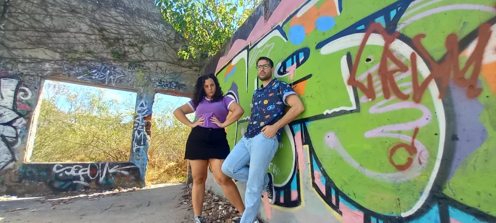 loca la calandria: "Fiore y Santi posan en un espacio urbano abandonado, junto a una pared con grafitis. La mujer lleva una camiseta morada y falda negra, mientras que el hombre viste una camisa estampada y jeans. La escena está iluminada por la luz del día, destacando el contraste entre la vegetación que se cuela por las ventanas sin vidrio y los coloridos grafitis en la pared.
