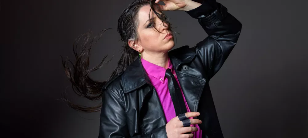 Lucy con campera de cuero negra y camisa fucsia con corbata, posando con el cabello suelto y la mano en la cabeza.