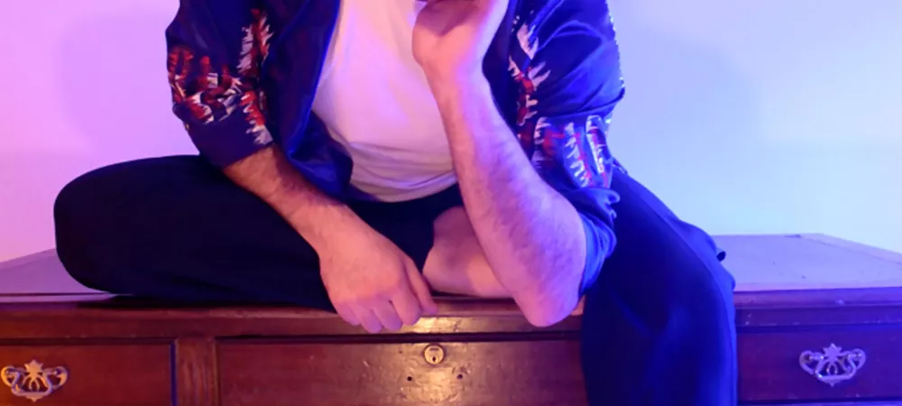 Manu Elle, sentado sobre un escritorio de madera con una pata cayendo delante y sosteniendo su cara con la mano, vestido con una remera blanco y una bata azul pintado con rojo y blanco. Hay luces celestes y rosas que impactan sobre una pared blanca de fondo