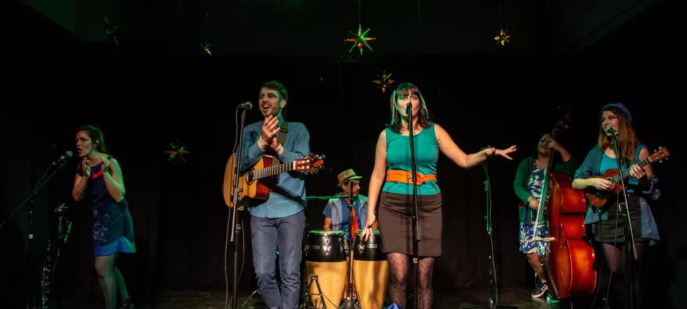 Se visualiza a la banda tocando y cantando. Se visualizan seis personas con instrumentos como: contrabajo, percusión, guitara, ukelele. Otros cantando y haciendo palmas.