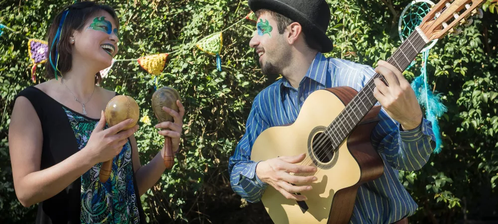 Si visualiza dos integrantes de la banda, una mujer sosteniendo dos maracas, un hombre sosteniendo una guitarra, que se miran uno al otro. 