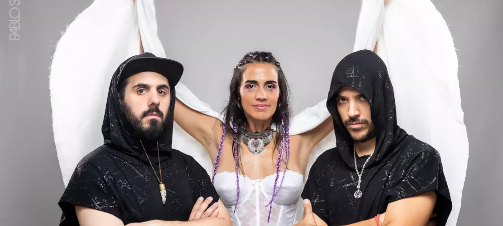 Trio de musicos, dos hombres, una mujer al medio con una capa que hace las veces de alas