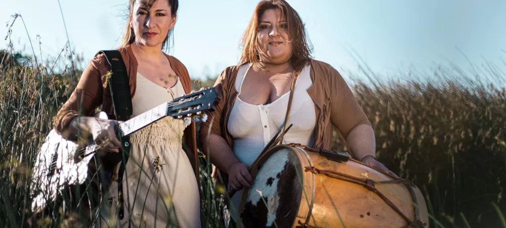 Mariel y Noelia estan con guitarra y bombo en mano entre juncos junto al rio